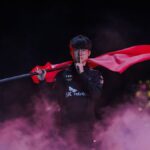 Faker, a League of Legends player, stands holding a finger to his mouth while holding a red T1 flag behind his back as smoke billows around him.